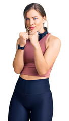 Beautiful caucasian young woman wearing gym clothes and using headphones ready to fight with fist defense gesture, angry and upset face, afraid of problem