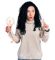 Young hispanic woman with curly hair holding small wooden manikin amazed and surprised looking up and pointing with fingers and raised arms.