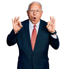 Senior man with grey hair wearing business suit and glasses looking surprised and shocked doing ok approval symbol with fingers. crazy expression