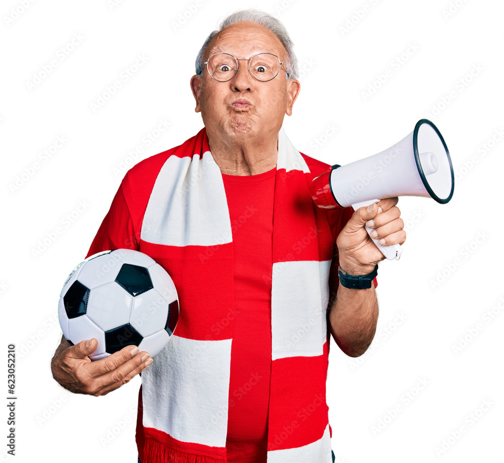 Wall mural Senior man with grey hair football hooligan holding ball and using megaphone puffing cheeks with funny face. mouth inflated with air, catching air.