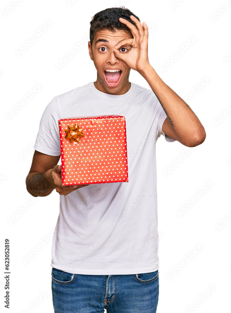 Wall mural Young handsome african american man holding gift smiling happy doing ok sign with hand on eye looking through fingers