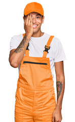 Young handsome african american man wearing handyman uniform covering one eye with hand, confident smile on face and surprise emotion.