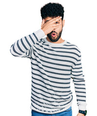 Young arab man with beard wearing casual striped sweater peeking in shock covering face and eyes with hand, looking through fingers with embarrassed expression.