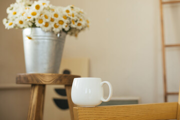 White mug with green tea and bouquet of chamomiles on wooden table in cozy living room.