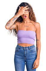 Young hispanic woman with tattoo wearing casual clothes peeking in shock covering face and eyes...