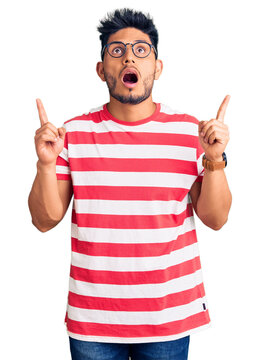 Handsome latin american young man wearing casual clothes and glasses amazed and surprised looking up and pointing with fingers and raised arms.