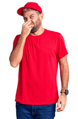 Young handsome blond man wearing t-shirt and cap smelling something stinky and disgusting,...
