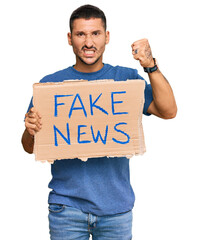 Handsome man with tattoos holding fake news banner annoyed and frustrated shouting with anger,...