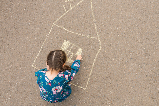 Junior school girl draws windows and curtains of house with white chalk on asphalt. Girl in flowery dress enjoys drawing house on asphalt