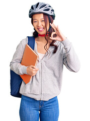 Young beautiful chinese girl wearing student backpack and bike helmet holding book doing ok sign with fingers, smiling friendly gesturing excellent symbol