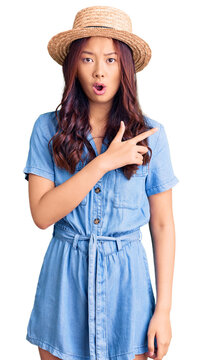 Young beautiful chinese girl wearing summer hat surprised pointing with finger to the side, open mouth amazed expression.