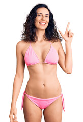 Young beautiful hispanic woman wearing bikini with a big smile on face, pointing with hand finger to the side looking at the camera. - obrazy, fototapety, plakaty