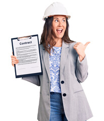 Young beautiful brunette woman wearing architect hardhat holding contract pointing thumb up to the side smiling happy with open mouth