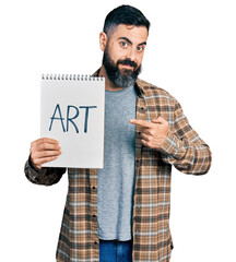 Hispanic man with beard holding art notebook smiling happy pointing with hand and finger