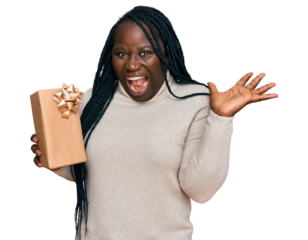 Young black woman with braids holding gift celebrating victory with happy smile and winner expression with raised hands
