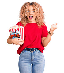 Young blonde woman with curly hair eating popcorn screaming proud, celebrating victory and success...