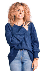 Young blonde woman with curly hair wearing casual winter sweater pointing aside worried and nervous with forefinger, concerned and surprised expression