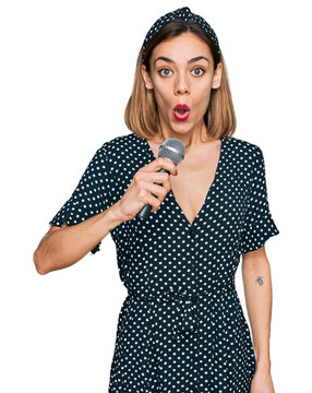 Young blonde girl singing song using microphone scared and amazed with open mouth for surprise, disbelief face