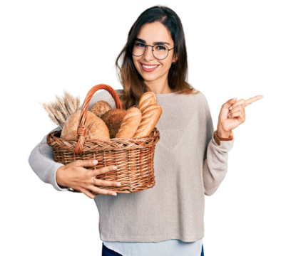 Beautiful hispanic woman holding wicker basket with bread smiling happy pointing with hand and finger to the side