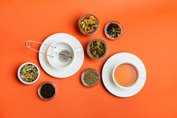 Obraz na płótnie Canvas White ceramic cup of tasty hot tea, empty cup and tea strainer, assortments tea leaves in small glass jars and bowls from above on orange background. Beverage for morning. Breakfast tea.