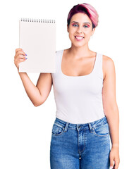 Young beautiful woman with pink hair holding notebook looking positive and happy standing and smiling with a confident smile showing teeth