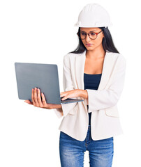 Young beautiful latin girl wearing architect hardhat holding laptop thinking attitude and sober expression looking self confident