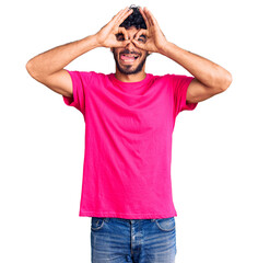 Handsome young man with curly hair and bear wearing casual pink tshirt doing ok gesture like binoculars sticking tongue out, eyes looking through fingers. crazy expression.