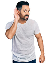 Young hispanic man with beard wearing casual grey t shirt smiling with hand over ear listening and hearing to rumor or gossip. deafness concept.
