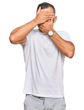 Middle age handsome man wearing casual white tshirt covering eyes and mouth with hands, surprised and shocked. hiding emotion