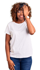 Young african american woman wearing casual white tshirt covering one eye with hand, confident smile on face and surprise emotion.