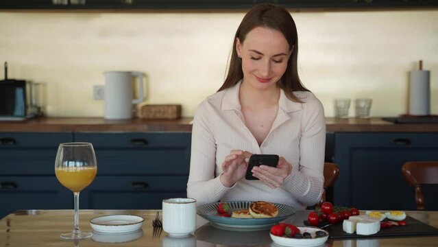 Happy woman uses a smartphone in the kitchen during a healthy breakfast in the morning, enjoys the weekend at home