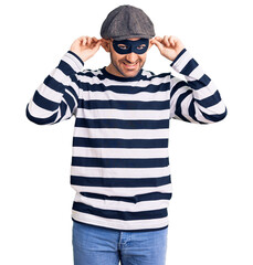 Young handsome man wearing burglar mask smiling pulling ears with fingers, funny gesture. audition problem