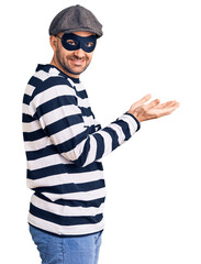 Young handsome man wearing burglar mask pointing aside with hands open palms showing copy space, presenting advertisement smiling excited happy