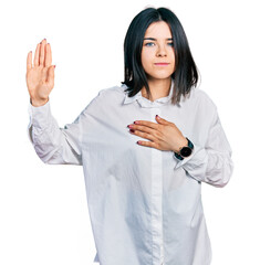 Fototapeta premium Young brunette woman with blue eyes wearing oversize white shirt swearing with hand on chest and open palm, making a loyalty promise oath
