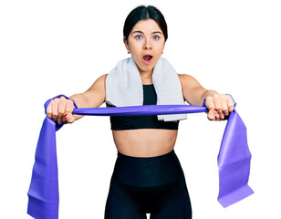 Young brunette woman with blue eyes training arm resistance with elastic arm bands afraid and shocked with surprise and amazed expression, fear and excited face.