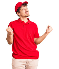 Young handsome man with curly hair wearing delivery uniform very happy and excited doing winner gesture with arms raised, smiling and screaming for success. celebration concept.