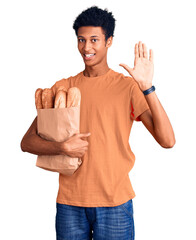 Young african american man holding paper bag with bread waiving saying hello happy and smiling, friendly welcome gesture
