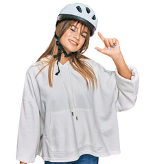 Teenager caucasian girl wearing bike helmet smiling and confident gesturing with hand doing small size sign with fingers looking and the camera. measure concept.