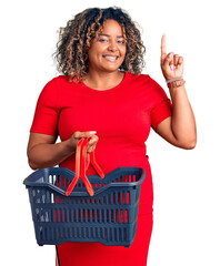 Young african american plus size woman holding supermarket shopping basket surprised with an idea...