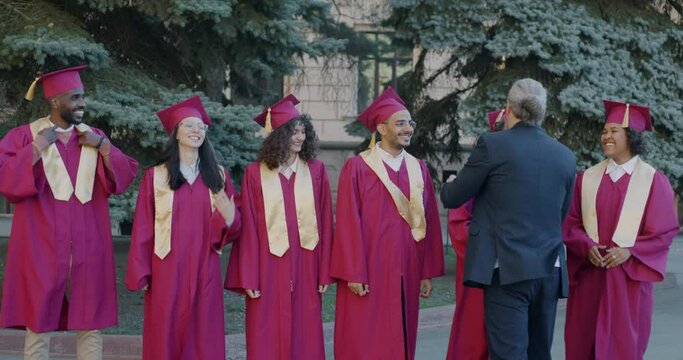 University dean congratulating group of students in graduation gowns and hats hugging youth clapping hands outdoors on campus. Education and achievement concept.