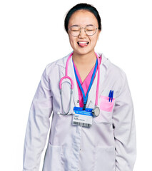 Young chinese woman wearing doctor uniform and stethoscope sticking tongue out happy with funny expression. emotion concept.