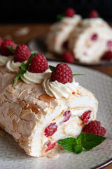 Side view of a meringue roll with raspberries and almond petals, decorated with cream, raspberries and mint leaves, on a dark wooden background. Vertical