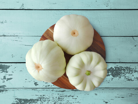 Three whole fresh pattypan squashes on round wooden cutting board on blue rough plank table surface top view      
