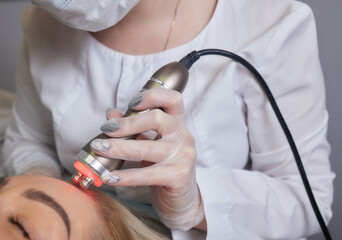 A cosmetologist in rubber gloves does the rf-lifting procedure on the face. In the background, the...