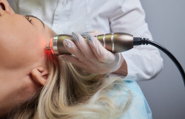 A cosmetologist in rubber gloves does the rf-lifting procedure on the face. In the background, the...