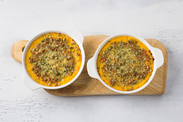 Homemade pumpkin casserole or gratin with cheese and pumpkin seeds in two ceramic tins on a light gray background