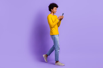 Full length photo of satisfied guy wear stylish yellow pullover look at smartphone walk empty space isolated on violet color background