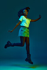 Portrait of cheerful, happy, young african woman in casual clothes posing, jumping with smile against cyan, blue studio background in neon light. Concept of emotions, youth, fashion, lifestyle, ad