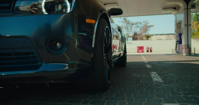 Close-up shot: a beautiful glossy dark gray convertible pulls into a gas station. The car is approaching the camera, view from below