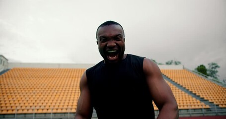A Black man in a black sports uniform poses and uses a battle cry in front of a stadium with yellow chairs in the stands